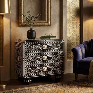 May include: A black and white floral patterned chest of drawers with three drawers and white flower-shaped knobs. The chest is set against a neutral-toned wall, with a gold-framed mirror and a blue armchair in the background.