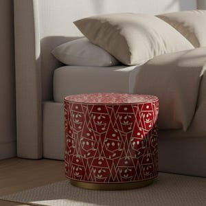 Könnte beinhalten: Ein rot-weiß gemusterter Beistelltisch mit goldenem Sockel. Der zylindrische Tisch steht auf einem neutralfarbenen Teppich in der Nähe eines Bettes mit hellem Kopfteil, Kissen und Bettwäsche. Das Design zeigt geometrische Formen und florale Elemente.