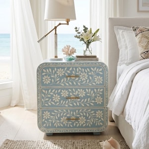 Pode incluir: Uma mesa de cabeceira azul clara com três gavetas, adornada com um padrão floral branco. A mesa tem puxadores de latão e está colocada ao lado de uma cama com roupa de cama branca. Uma lâmpada e objetos decorativos estão em cima.