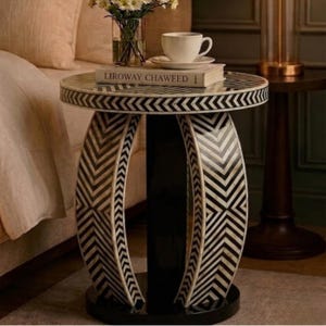 May include: A small side table with a round top featuring a black and white chevron design. The table has a black base and central support. A book, a cup of coffee, and flowers are on the table.