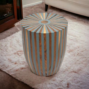 Puede incluir: Una mesa auxiliar cilíndrica de madera con un diseño de rayos de sol en la parte superior. La mesa presenta rayas verticales alternas de color azul claro y madera natural, con detalles dorados. La mesa mide aproximadamente 46 cm de altura.