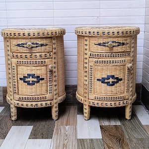 May include: Two matching wicker bedside tables. Each has a rounded top and three drawers. The drawers are accented with black and gold details. The tables are made of woven wicker and have four short legs.