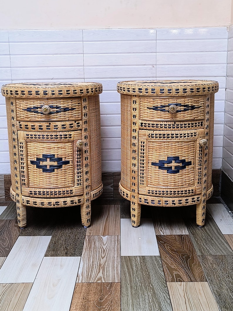 May include: Two matching wicker bedside tables. Each has a rounded top and three drawers. The drawers are accented with black and gold details. The tables are made of woven wicker and have four short legs.