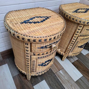 May include: Two woven wicker bedside tables. The tables are oval-shaped with a light brown colour and dark blue geometric designs. Each table has drawers and a decorative handle. The tables are on wooden legs.