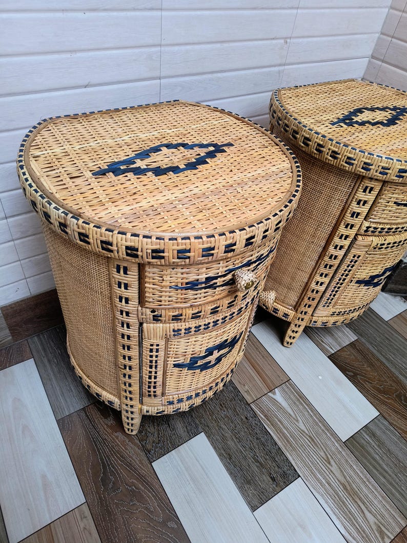 May include: Two woven wicker bedside tables. The tables are oval-shaped with a light brown colour and dark blue geometric designs. Each table has drawers and a decorative handle. The tables are on wooden legs.