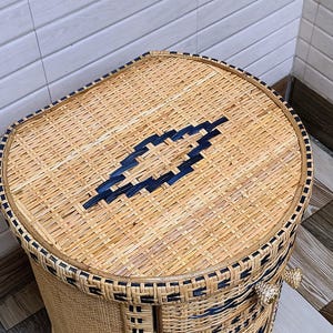 May include: Woven wicker storage basket with a rounded top and a rectangular body. The basket is made of natural-coloured wicker with dark blue accents and trim. The lid features a geometric design.