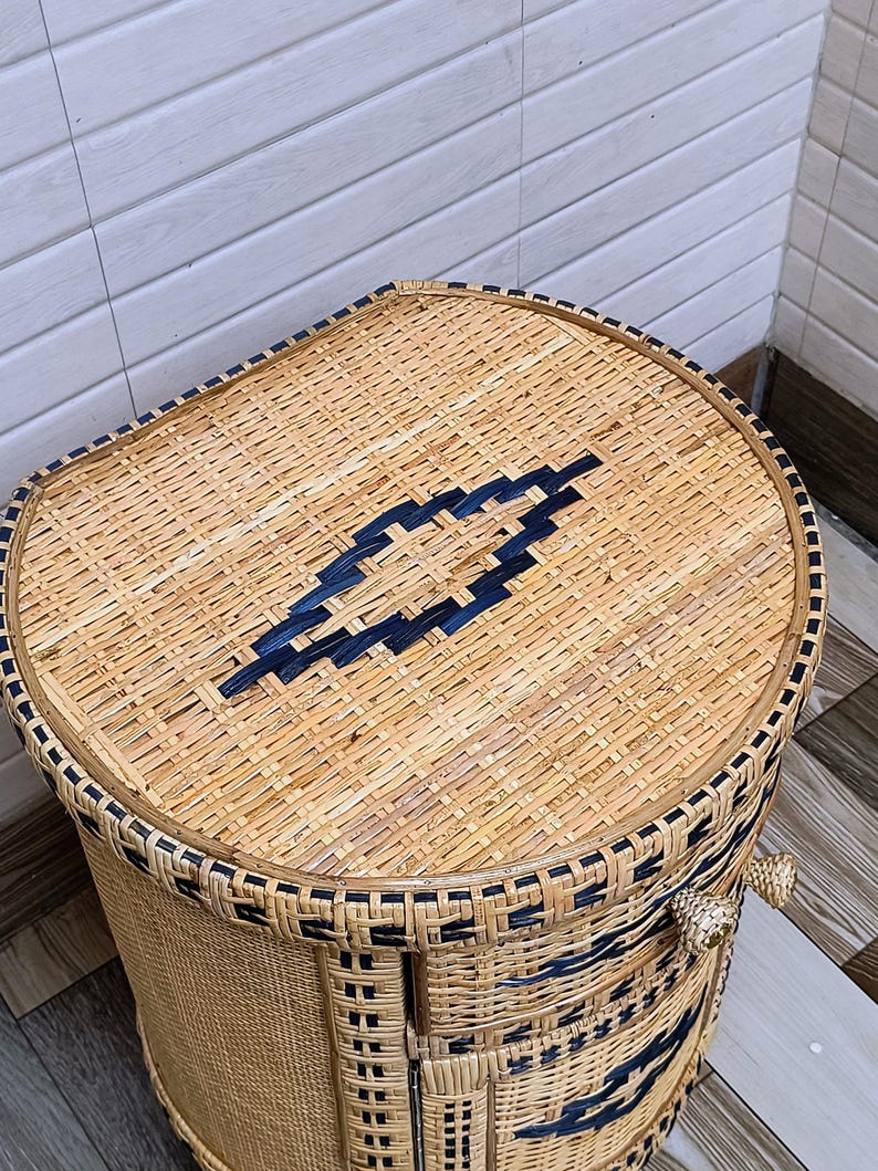 May include: Woven wicker storage basket with a rounded top and a rectangular body. The basket is made of natural-coloured wicker with dark blue accents and trim. The lid features a geometric design.