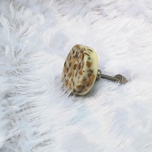 Puede incluir: Pomo decorativo para armario con base color crema y motas doradas. El pomo está sujeto a un tornillo y tuerca metálicos, sobre un fondo blanco y esponjoso. Este herraje es adecuado para cajones y armarios.