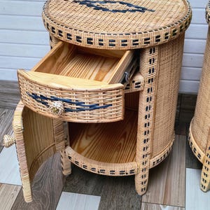 May include: Wicker bedside table with a round top and a drawer. The table is made of light brown wicker with black accents and a wooden interior. The drawer is open, revealing the interior. The table has a small door on the side.