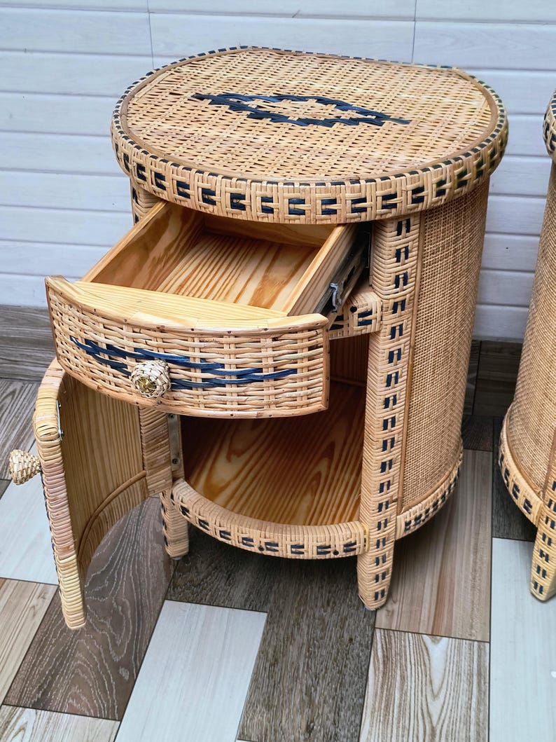 May include: Wicker bedside table with a round top and a drawer. The table is made of light brown wicker with black accents and a wooden interior. The drawer is open, revealing the interior. The table has a small door on the side.