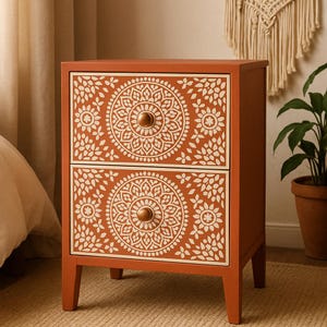 May include: A two-drawer nightstand in a warm terracotta color. The drawers feature a white, intricate mandala design and round, bronze-colored knobs. The nightstand has a rectangular shape and sits on four short legs.