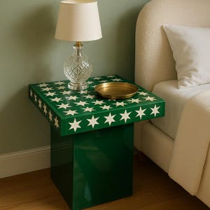 Puede incluir: Una mesita de noche verde con un patrón de estrellas blancas. Una lámpara de cristal transparente con una pantalla color crema y una pequeña bandeja dorada están sobre la mesa. La mesa está junto a una cama con una colcha blanca. La mesa mide unos 50 cm de alto.