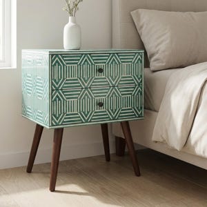 May include: A green and white geometric patterned bedside table with two drawers and dark brown legs. A white vase with flowers sits on top. The table is next to a bed with a beige headboard.