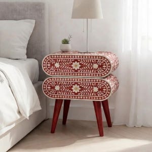 May include: A red and white floral patterned bedside table with a stacked oval design. The table has four tapered legs. A lamp and small plant sit on top. The table is next to a bed.
