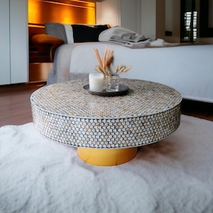 May include: A round, white and gold coffee table with a mosaic top made of small, iridescent shells. The table is sitting on a white rug in front of a bed.