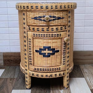 May include: A woven wicker bedside table with a rounded shape. The table has two drawers and a door, all with dark blue woven accents. The drawer and door pulls are gold-coloured. The table is supported by four short legs.