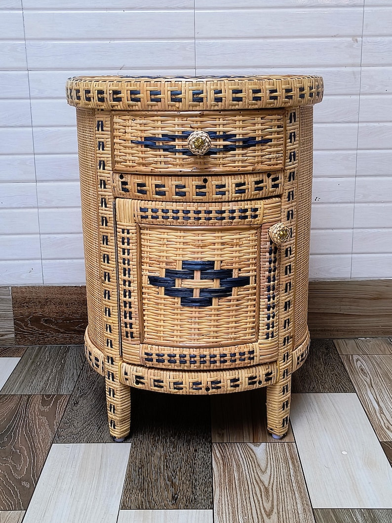 May include: A woven wicker bedside table with a rounded shape. The table has two drawers and a door, all with dark blue woven accents. The drawer and door pulls are gold-coloured. The table is supported by four short legs.
