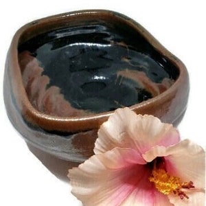 Puede incluir: Un pequeño cuenco cuadrado de cerámica marrón y negro con una flor en el lateral. El cuenco tiene un acabado esmaltado y un borde ligeramente irregular.