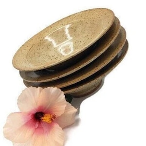 Puede incluir: Un juego de cuatro cuencos de cerámica marrón moteados apilados uno encima del otro. Los cuencos son hechos a mano y tienen un aspecto rústico. Una flor de hibisco rosa está en primer plano.