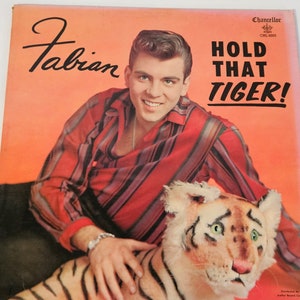Puede incluir: Una portada de disco de vinilo vintage con Fabian, un joven con cabello oscuro, vistiendo una camisa a rayas rojas y negras, sosteniendo un tigre de peluche. La portada es naranja y tiene el título "Hold That Tiger!" en letras negras.