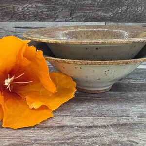 May include: Two ceramic bowls with a speckled, earthy tone are stacked, showcasing a rustic aesthetic. A vibrant orange flower with a white center is positioned to the left, adding a pop of color. The bowls have a wide rim and a shallow depth.