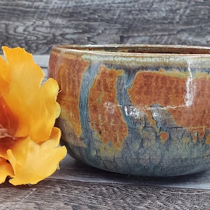 Puede incluir: Un cuenco de cerámica hecho a mano con un esmalte marrón, naranja y azul. El cuenco tiene una forma redondeada y un borde ancho.