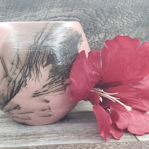 May include: A small, round, pink ceramic vase with black abstract designs. The vase is sitting on a wooden surface next to a red flower.