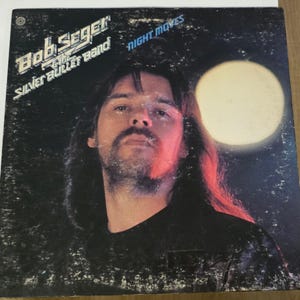 May include: Album cover for Bob Seger & The Silver Bullet Band's "Night Moves." The album art features a portrait of a man with long hair and a mustache, set against a dark background with a large, glowing orb. The band's name and album title are printed in white.