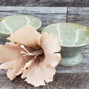 May include: Two green ceramic bowls with a speckled glaze. The bowls have a wide rim and a narrow base. A light brown flower is in the center of the image.