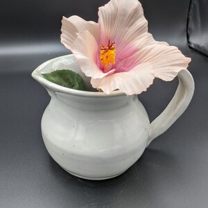 May include: A white ceramic pitcher with a wide mouth and a handle. A pink hibiscus flower with a yellow center is placed inside the pitcher.