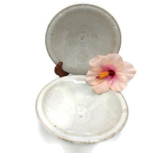 Puede incluir: Dos cuencos de cerámica blanca con un esmalte moteado. Los cuencos están apilados uno encima del otro y una flor de hibisco rosa descansa sobre el cuenco superior.