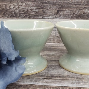 May include: Two light blue ceramic cups with a flared rim and a pedestal base. The cups are sitting on a wooden surface.