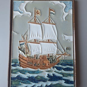 Puede incluir: Una placa de pared de cerámica que representa un barco de vela de tres mástiles con velas blancas, casco marrón y olas azules y blancas. El barco tiene banderas con un león y una cruz.