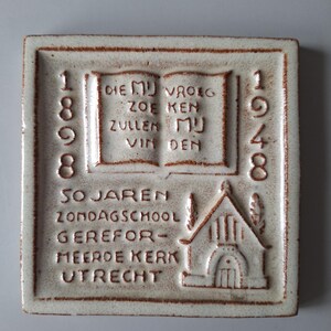 Può includere: Piastrella quadrata in ceramica bianco sporco con testo e immagini in rilievo. La piastrella presenta gli anni 1898 e 1948, testo in olandese e un'illustrazione di una chiesa. Il testo include "SOJAREN ZONDAGSCHOOL GEREFORMEERDE KERK UTRECHT."
