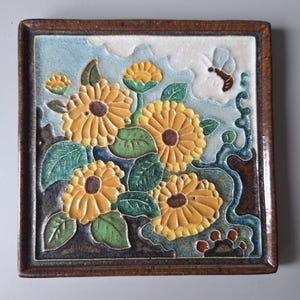 Op de afbeelding: Vierkante keramische tegel met een verhoogd bloemmotief. De tegel heeft gele zonnebloemen met bruine centra, groene bladeren en een blauwe luchtachtergrond. Een bruin houten frame omringt de tegel en een bij is zichtbaar bij de bloemen.