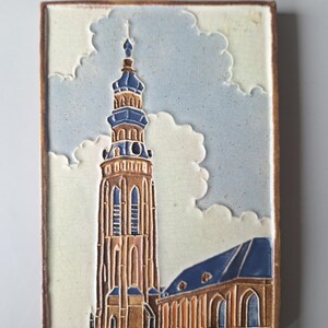 Może przedstawiać: Prostokątna płytka ceramiczna z szczegółową ilustracją wysokiej wieży kościelnej w niebiesko-brązowej kolorystyce, na tle zachmurzonego nieba. Płytka ma brązową obwódkę i napis "MIDDELBURG-LANGE JAN" na dole.