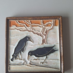May include: Square ceramic tile featuring a nature scene with two black crows and a bare tree. The tile has a brown border and a background with orange and white tones. The crows are depicted in a stylized manner.
