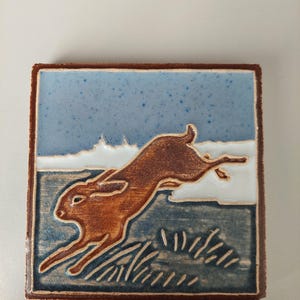 May include: A square ceramic tile featuring a relief design of a brown hare leaping across a field. The background consists of a speckled light blue sky above a dark blue landscape with white accents. The tile has a dark brown border.