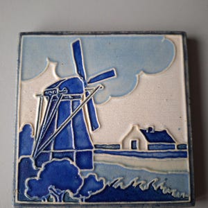 Puede incluir: Un azulejo de cerámica azul y blanco con un molino de viento y una pequeña casa en el fondo. El azulejo tiene un esmalte azul y blanco y está decorado con un diseño estilizado.