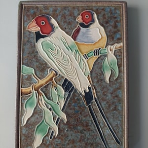 May include: A rectangular ceramic tile featuring a relief design of two Gouldian finches perched on a branch. The birds have red faces, black throats, and white, green, and yellow plumage. The background is a textured, mottled blue-brown finish.