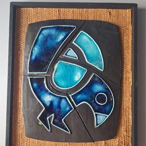 May include: A framed ceramic wall art piece featuring a stylized bird design. The bird is composed of blue and turquoise glazed ceramic tiles set against a dark gray background. The frame is black with a woven straw-like border.