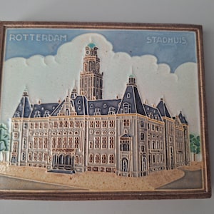 May include: A rectangular ceramic tile featuring a relief depiction of the Rotterdam City Hall, labeled ROTTERDAM STADHUIS. The building is rendered in shades of gray and blue against a light blue sky with white clouds, framed by a dark brown border.