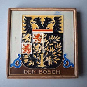 Op de afbeelding: Een ingelijste keramische tegel met een wapenschild met een boom, leeuwen en vogels. De tegel is bruin met blauwe en gele accenten. De tekst "DEN BOSCH" is onder het wapenschild gedrukt.