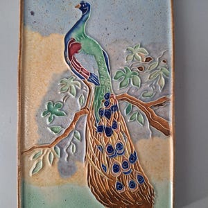 May include: A rectangular ceramic wall tile featuring a relief design of a peacock perched on a brown tree branch. The peacock displays green, blue, and red plumage with detailed tail feathers. The background is a mottled mix of light blue, tan, and green.