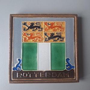 Może przedstawiać: Kwadratowa płytka ceramiczna przedstawiająca herb Rotterdamu. Wzór zawiera cztery lwy w kolorze czarnym i pomarańczowym na złotym tle nad polem w zielono-białe pionowe pasy. Słowo ROTTERDAM jest wytłoczone na brązowej podstawie płytki.