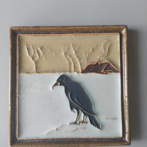 May include: Square ceramic tile with a dark brown frame. The tile features a black crow standing on a white surface, with a brown and tan landscape background. The crow is the focal point of the tile.