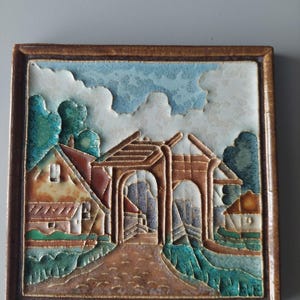 May include: A square ceramic wall tile featuring a relief landscape scene of a village with a stone archway, houses, and trees. The design uses a palette of brown, teal, and cream glazes, framed by a raised brown border with a textured finish.