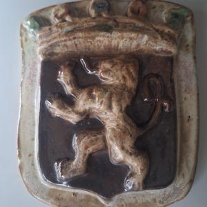 Może przedstawiać: Ceramiczna tablica ścienna w kształcie tarczy, przedstawiająca wypukły relief lwa w pozycji wspiętej, pokryty ciemnobrązowym szkliwem. Krawędź oraz korona u góry wykończone są jaśniejszym, nakrapianym beżowym szkliwem z subtelnymi zielonymi akcentami.