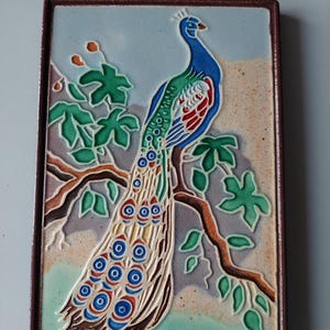 May include: A rectangular ceramic tile featuring a raised relief design of a peacock perched on a brown tree branch. The peacock has a blue head and neck, green and red wing details, and a long tail with blue and red eye-patterned feathers.