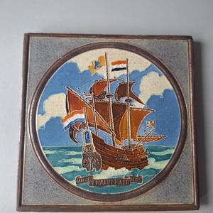 Puede incluir: Un azulejo de cerámica cuadrado con un diseño circular que presenta un barco de vela marrón con velas blancas y detalles naranjas. El barco navega en un mar azul con nubes blancas y un cielo azul. El azulejo tiene un borde marrón y el texto "MARE DE NAUWE MAER 1600" en una pancarta.